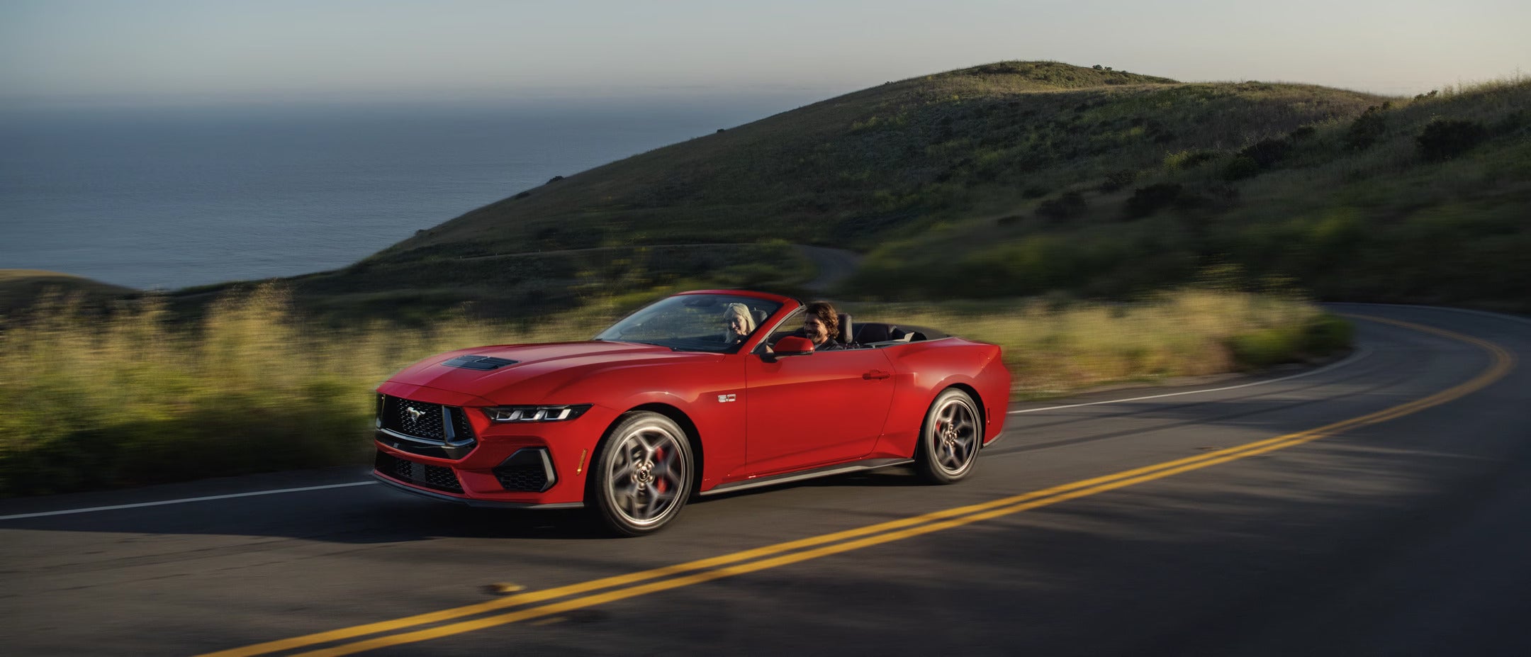 Side view of a red 2025 Ford Mustang convertible driving along a coastal road.