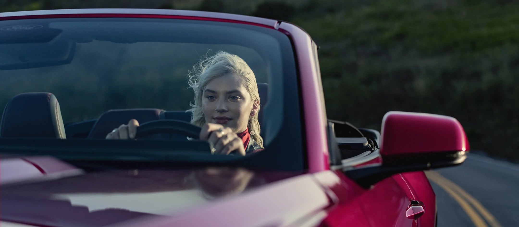 Front view of a driver in a red 2025 Ford Mustang convertible driving along a scenic road